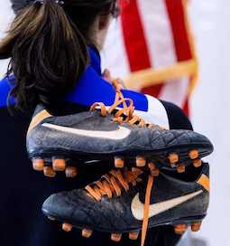 A pair of cleated shoes held over the shoulder of a woman in front of an American flag.
