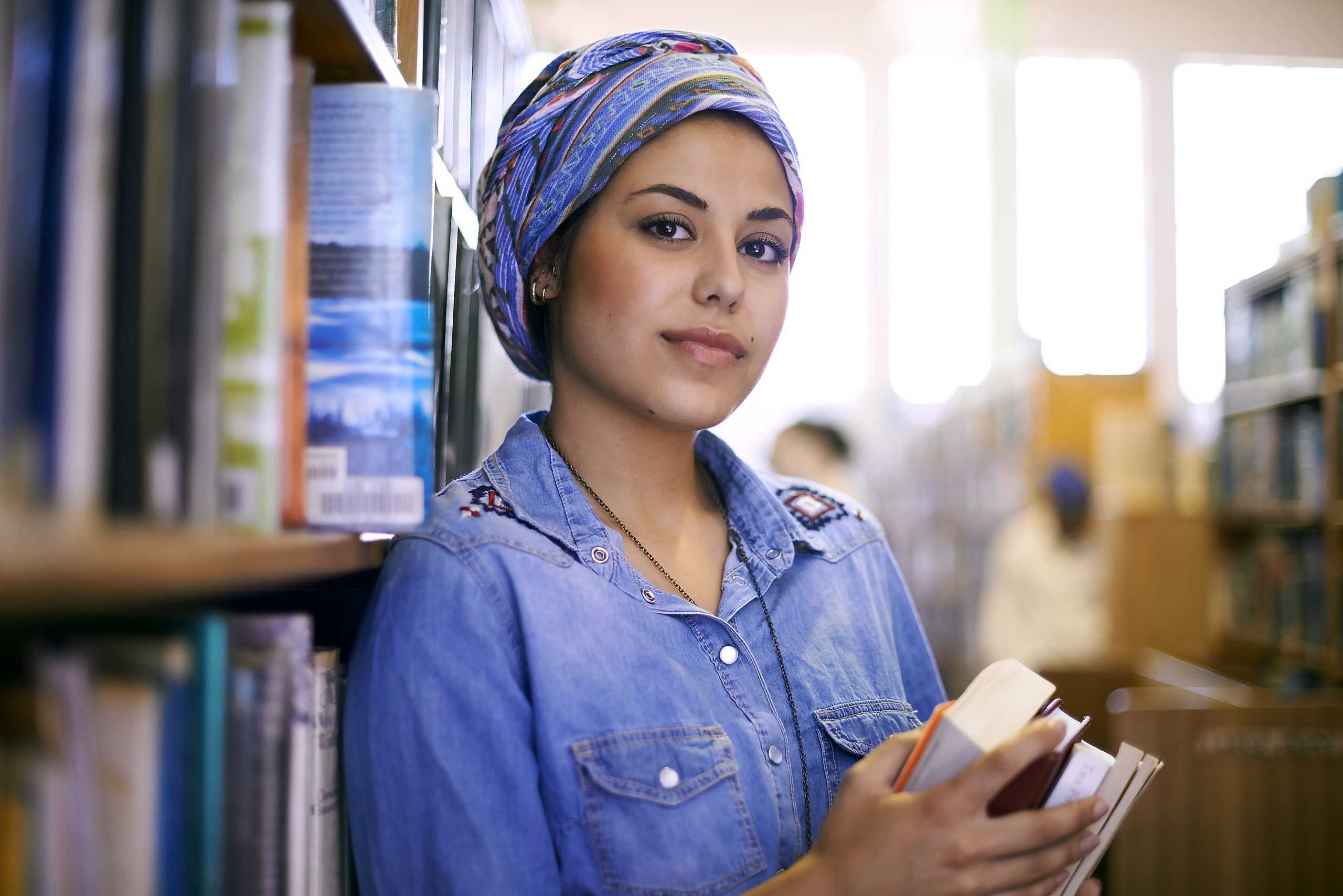A woman with medium-light skin wearing a head scarf holding a stack of books in a library.