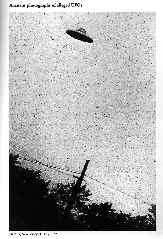 A black and white photograph of a small UFO flying atop the silhouette of trees and a powerline.