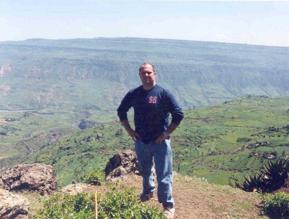 A color photograph of Gregg standing on top of a cliff overseeing a lush green valley.