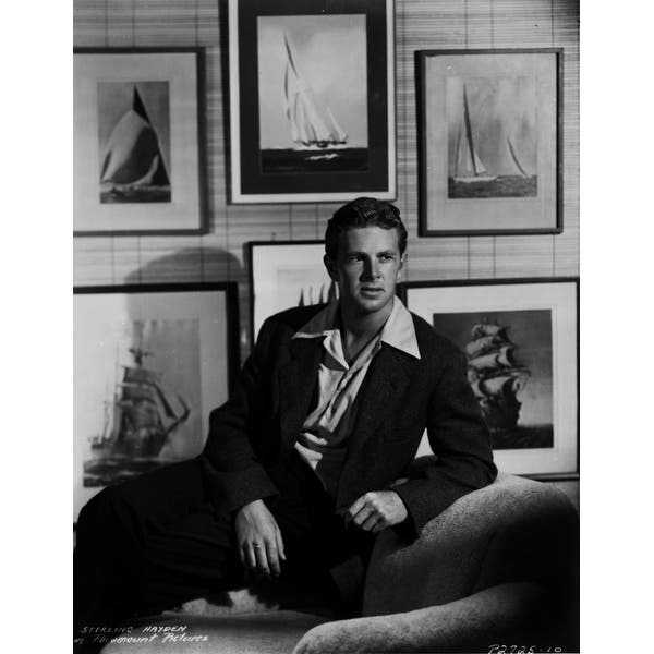 black and white photo of man posed, lounging on a sofa with photographs on wall behind him.