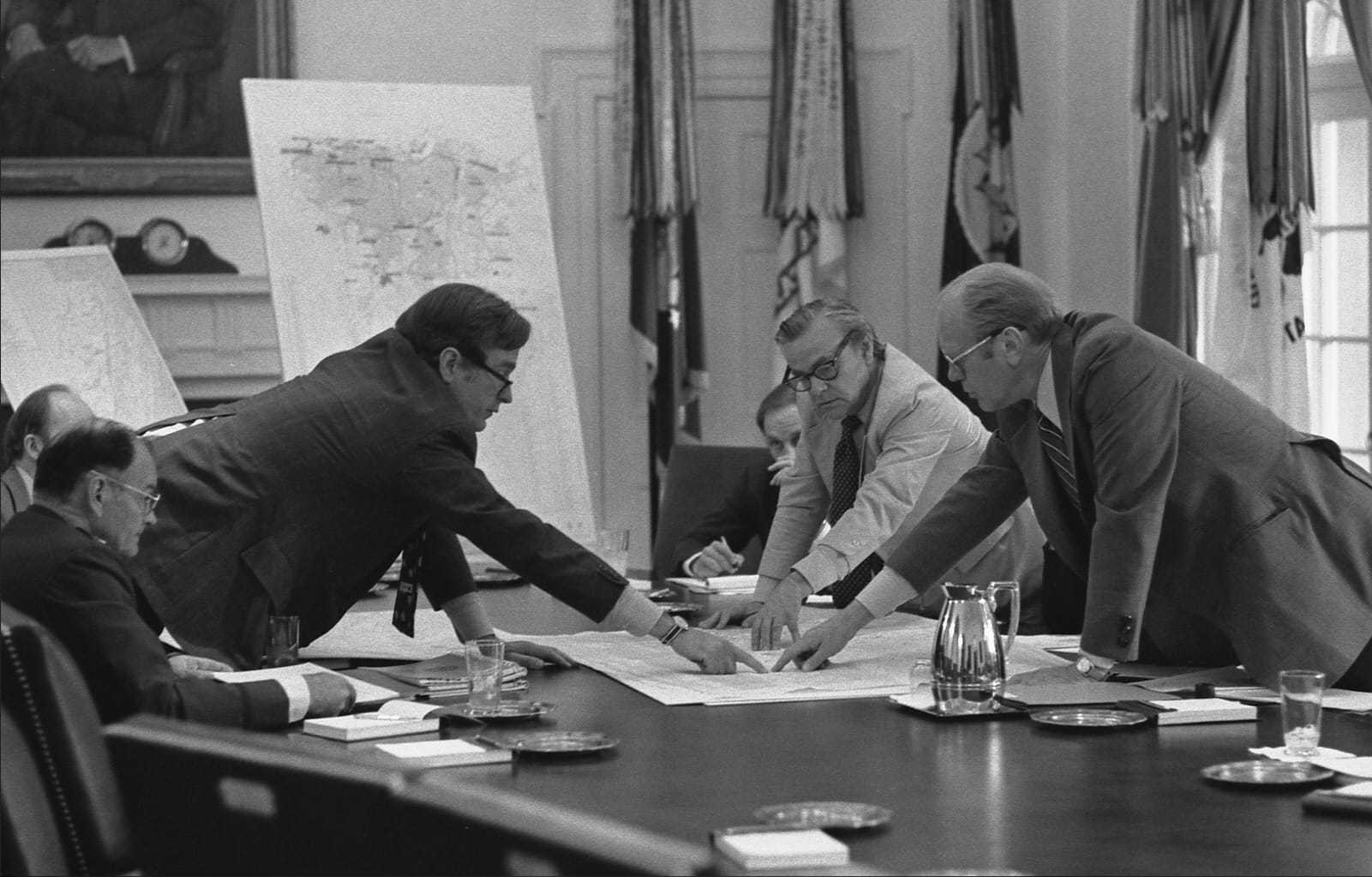 DCI George Bush points at a paper on a table with President Ford and three others around the table.