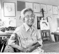 black and white photo of older woman sitting, smiling at camera in what looks like an artist's studio