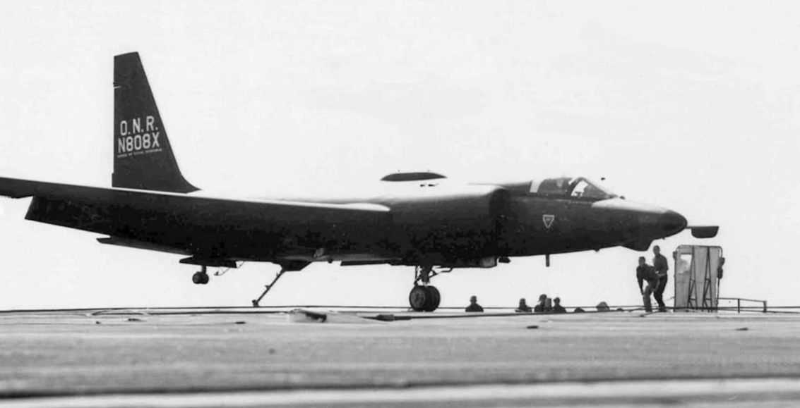 A black and white image of an aircraft on a tarmac with two people standing under its nose.
