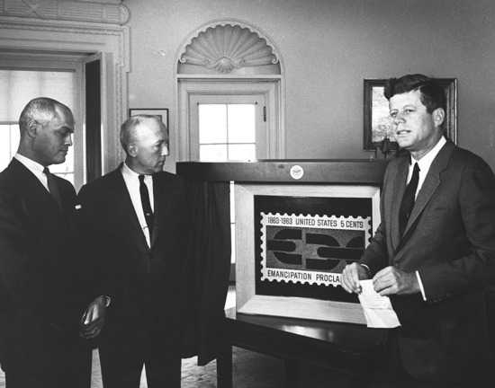 black and white image of three men, including President Kennedy, standing around a large photograph of a postage stamp.