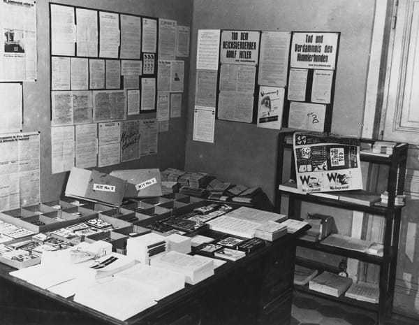 An OSS printshop with propaganda and leaflets in piles on the desk and on the walls.