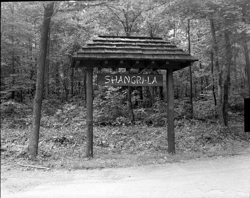1950s black and white photo of wooden sign on the side of the road with a forest in the background. Sign reads Shangri-La.