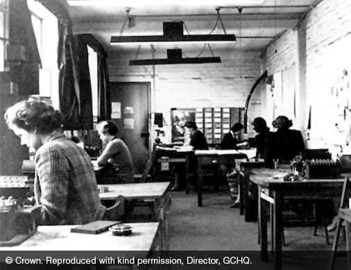 A black and white photo of mathematicians in a room with desks, working on the ENIGMA machine.