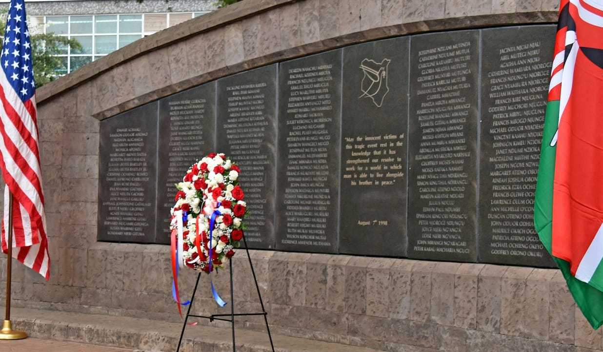 A memorial plaque with names inscribed in gold, with a wreath boquet of flowers in front of it and the American and Kenyan flags at its sides.