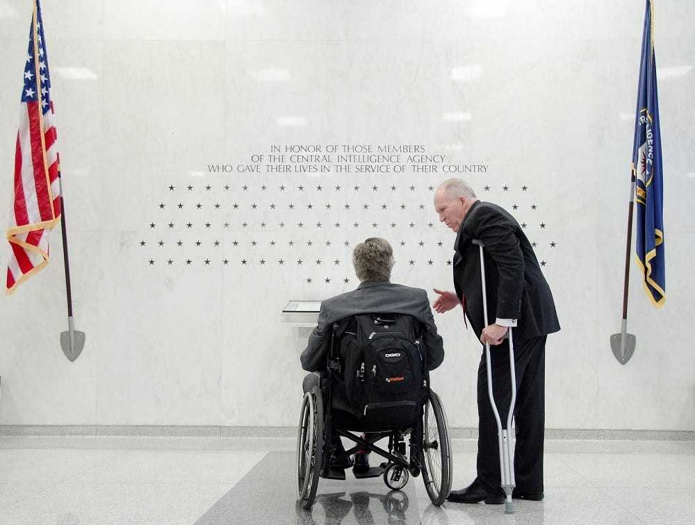 Bush, in a wheelchair, and Director John Brennan, with a crutch under his left armpit, looking at a memorial inscribed on the sheen white wall inside the entrance of the CIA Headquarters Building.