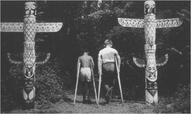 1930s black and white photo showing two student-made totem poles with two young students on crutches in the center, facing with their backs to the camera walking down a path.