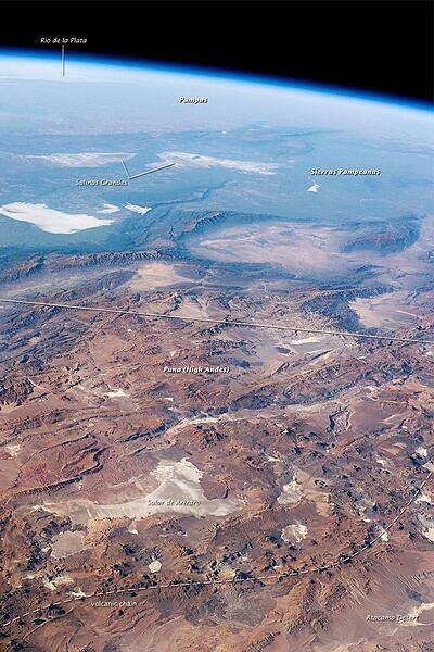 This panorama looking southeast across the South American continent was taken from the International Space Station almost directly over the Atacama Desert near Chile's Pacific coast. The high plains (3000-5000 m, 13,000-19,000 ft) of the Andes Mountains, also known as the Puna, appear in the foreground, with a line of young volcanoes (dashed line) facing the much lower Atacama Desert (1000-2000 m elevation). Several salt-crusted dry lakes (known as salars in Spanish) occupy the basins between major thrust faults in the Puna. Salar de Arizaro (foreground) is the largest of the dry lakes in this view. The Atlantic Ocean coastline, where Argentina's capital city of Buenos Aires sits along the Río de la Plata, is dimly visible at image top left. Near image center, the transition (solid line) between two distinct geological zones, the Puna and the Sierras Pampeanas, creates a striking landscape contrast. Compared to the Puna, the Sierras Pampeanas mountains are lower in elevation and have fewer young volcanoes. Sharp-crested ridges are separated by wide, low valleys in this region. The Salinas Grandes - ephemeral shallow salt lakes - occupies one of these valleys. The general color change from reds and browns in the foreground to blues and greens in the upper part of the image reflects the major climatic regions: the deserts of the Atacama and Puna versus the grassy plains of central Argentina, where rainfall is sufficient to promote lush prairie grass, known locally as the pampas. The Salinas Grandes mark an intermediate, semiarid region. Image courtesy of NASA.
