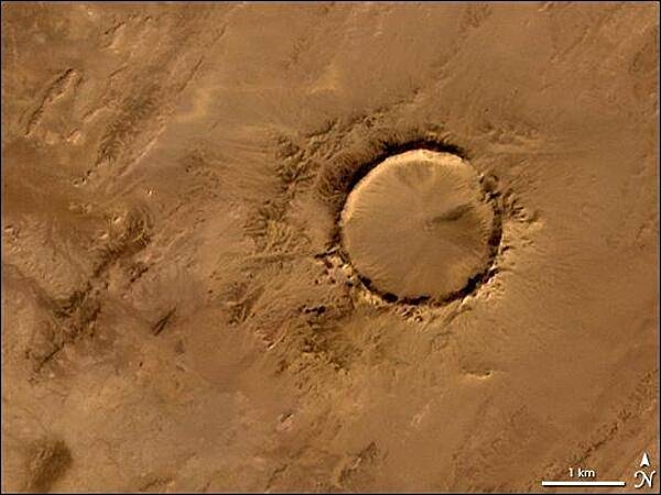 Deep in the Sahara lies a crater, a nearly a perfect circle that is 1.9 km (1.2 mi) wide, and sports a rim 100 m (330 ft) high. The Tenoumer crater sits in a vast plain of rocks so ancient they were deposited hundreds of millions of years before the first dinosaurs walked the Earth. Close examination of the structure has revealed that the crater's hardened "lava" was actually rock that had melted from a meteorite impact.
On this satellite image the crater's outline is unmistakable, yet it does not necessarily look like a crater; the light and shadows make it look more as if someone pressed a giant cookie cutter into the rock. In this image, the sunlight shines from the southeast (lower right), and the bright arc along the northwestern part of the crater is where the crater walls slope up to the rim. Around the perimeter, the relatively steep walls cast dark shadows. Although it resides in ancient rock, Tenoumer is geologically young, ranging in age between roughly 10,000 and 30,000 years old. Photo courtesy of NASA.