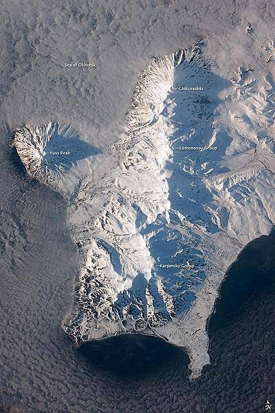 The Kuril Island chain is built from a line of volcanoes, an island arc, that extends from Russia's Kamchatka Peninsula to northern Japan. Island arcs form along an active boundary between two tectonic plates, with one being driven beneath the other (subduction). Magma generated by subduction feeds volcanoes - and eventually volcanic islands - over the subduction boundary. Paramushir Island in the northern Kurils is an example of a large island built by several volcanoes over geologic time. This astronaut photograph shows the southern end of Paramushir Island after a snowfall. The western slopes of the mountains are brightly illuminated, while the eastern slopes are in shadow.
Four major volcanic centers create this part of the island. Fuss Peak (image center left) is an isolated stratovolcano connected to the main island via an isthmus. Fuss Peak last erupted in 1854.
The southern tip of the island is occupied by the Karpinsky Group of three volcanic centers. A minor eruption of ash following an earthquake occurred on this part of the island in 1952.
The Lomonosov Group to the northeast (image center) includes four cinder cones and a lava dome.
The most recent volcanic activity on Paramushir Island occurred in 2008 at the Chikurachki cone located along the northern coastline of the island at image top center. The summit of this volcano, 1,816 m (5,958 ft) above sea level, is the highest on Paramushir Island.
Much of the Sea of Okhotsk visible in the image is covered with low clouds that often form around the islands in the Kuril chain. The clouds are generated by moisture-laden air passing over the cool sea/ocean water, and they typically wrap around the volcanic islands. Image courtesy of NASA.
