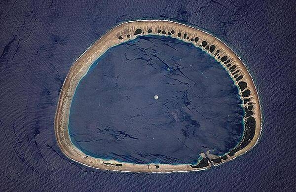Classically shaped Nukuoro Atoll is one of 607 islands that make up the Federated States of Micronesia. It is also a municipality of the state of Pohnpei, Federated States of Micronesia. The atoll has a population of only several hundred people. The total area, including the lagoon, is 40 sq km, with a land area of only 1.7 sq km, which is divided among more than 40 islets that lie on the northern, eastern, and southern sides of the lagoon. By far the largest islet is Nukuoro islet in the southeast, which is the center of population and the capital of the municipality. Vegetation is most prominent on the northeast and east portions of the atoll that face the dominant Easterly winds. Swells driven by these winds can be seen approaching from the east-southeast (right) and wrapping around the atoll to produce an interference pattern on the downwind side. Water in the lagoon is calm in comparison. A coral head appears dead center of the photo. Image courtesy of NASA.