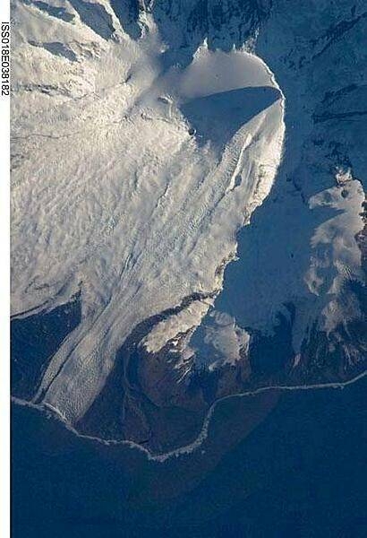 The northeastern slopes of Big Ben volcano on Heard Island are visible in shadow at the top right of this photo. Mawson Peak (small circle in the upper center of the image), sits within a breached caldera (collapsed and empty magma chamber beneath a volcano) on the southwestern side of the volcano - the shadow cast by Mawson Peak points directly to the crescent-shaped caldera rim. This image, taken during the Southern Hemisphere summer, also shows the dark brown volcanic rock that forms the island. The southwest-pointing tongue of ice is Gotley Glacier. Image credit: NASA.